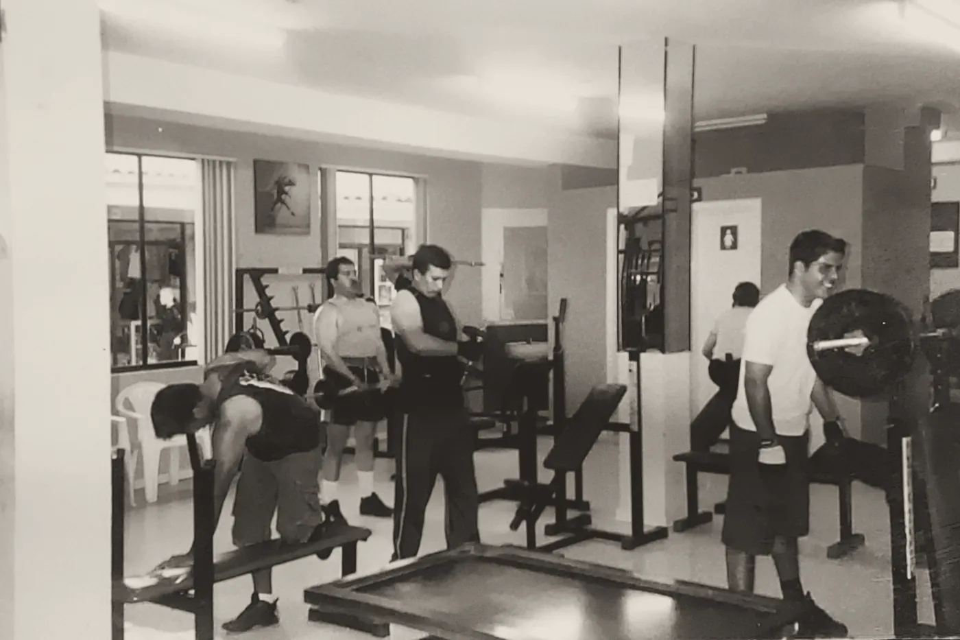 Socios entrenando con barras y bancos en una foto antigua del gimnasio.